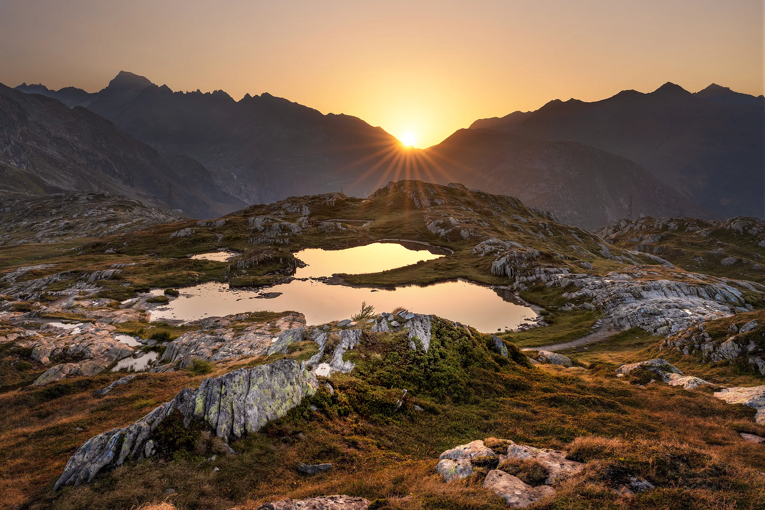 Sun setting behind a mountain range, with rocky terrain and small water bodies in the foreground.