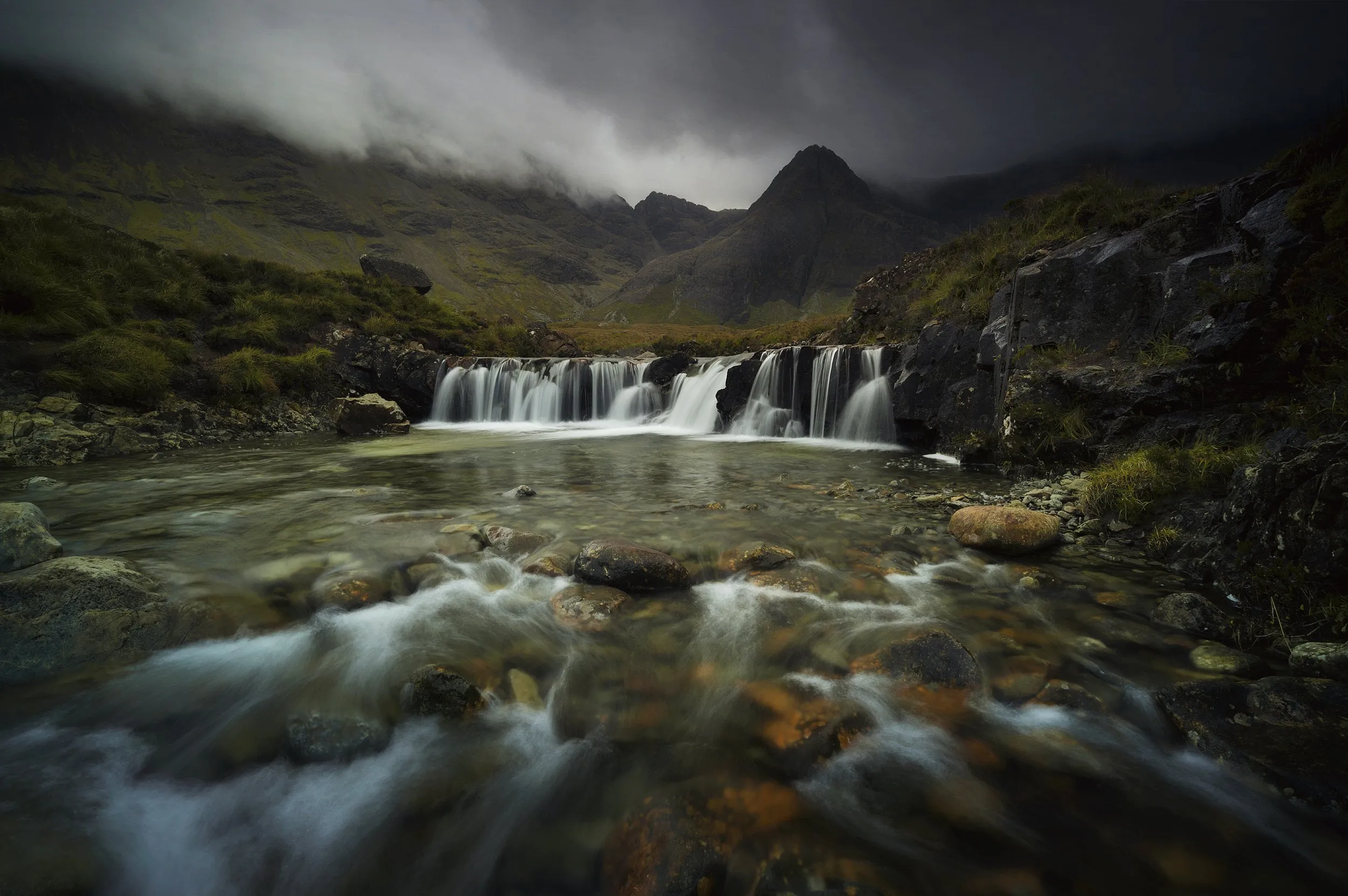 A waterfall feeding a rocky mountain stream under a clouded sky.