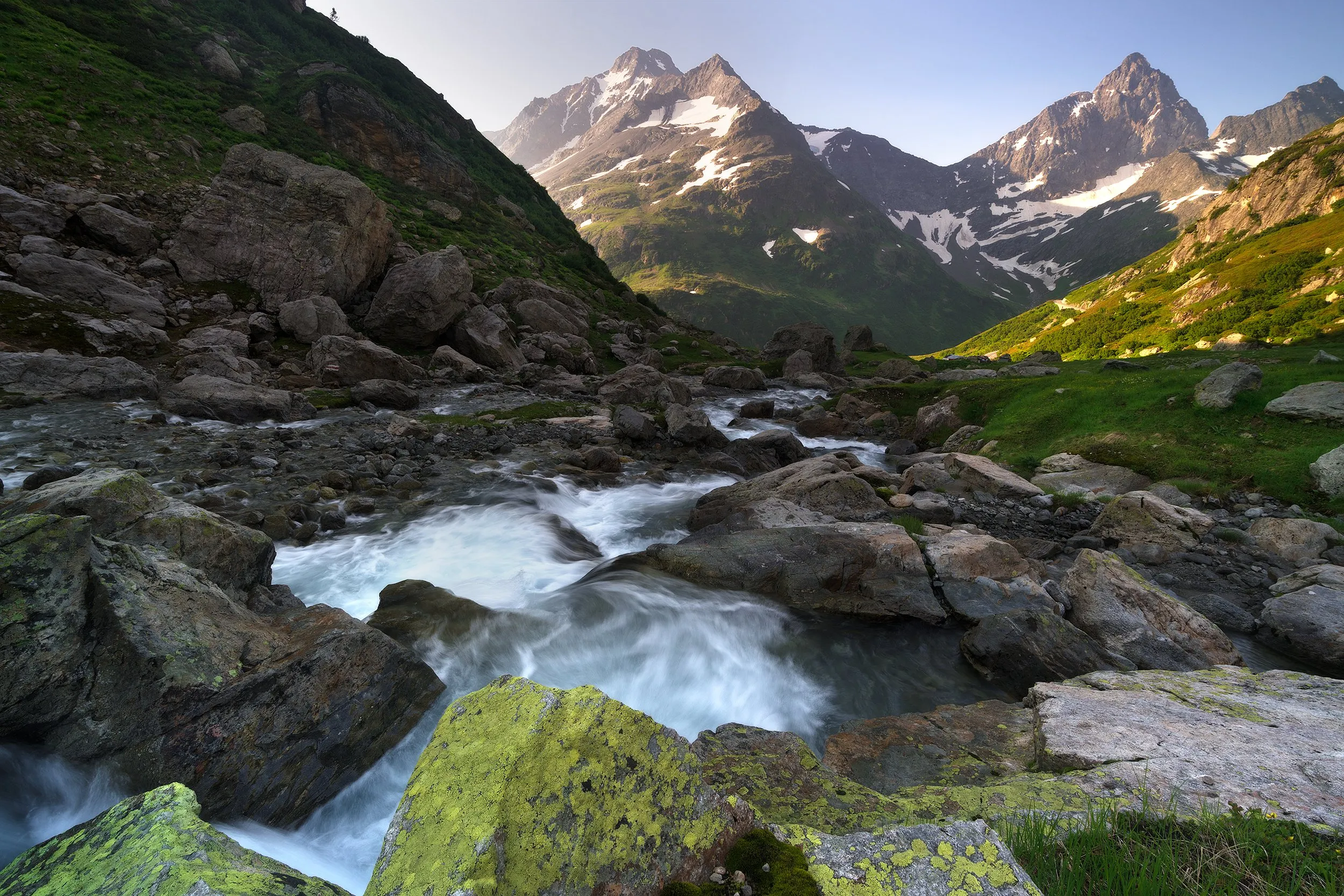 Mountain stream through a green valley framed by snow-capped peaks.