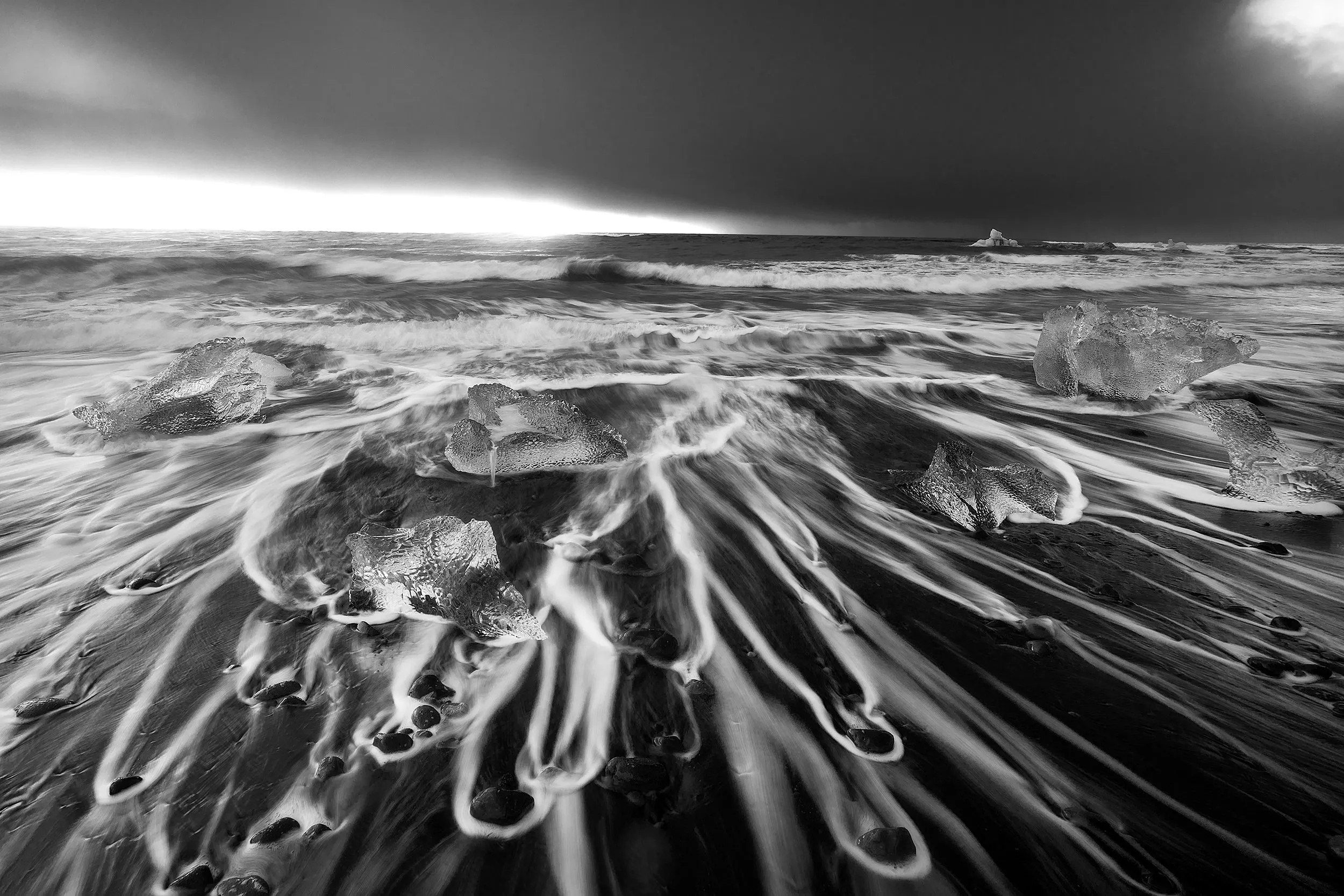 Waves breaking over an ice-strewn beach, in monochrome.