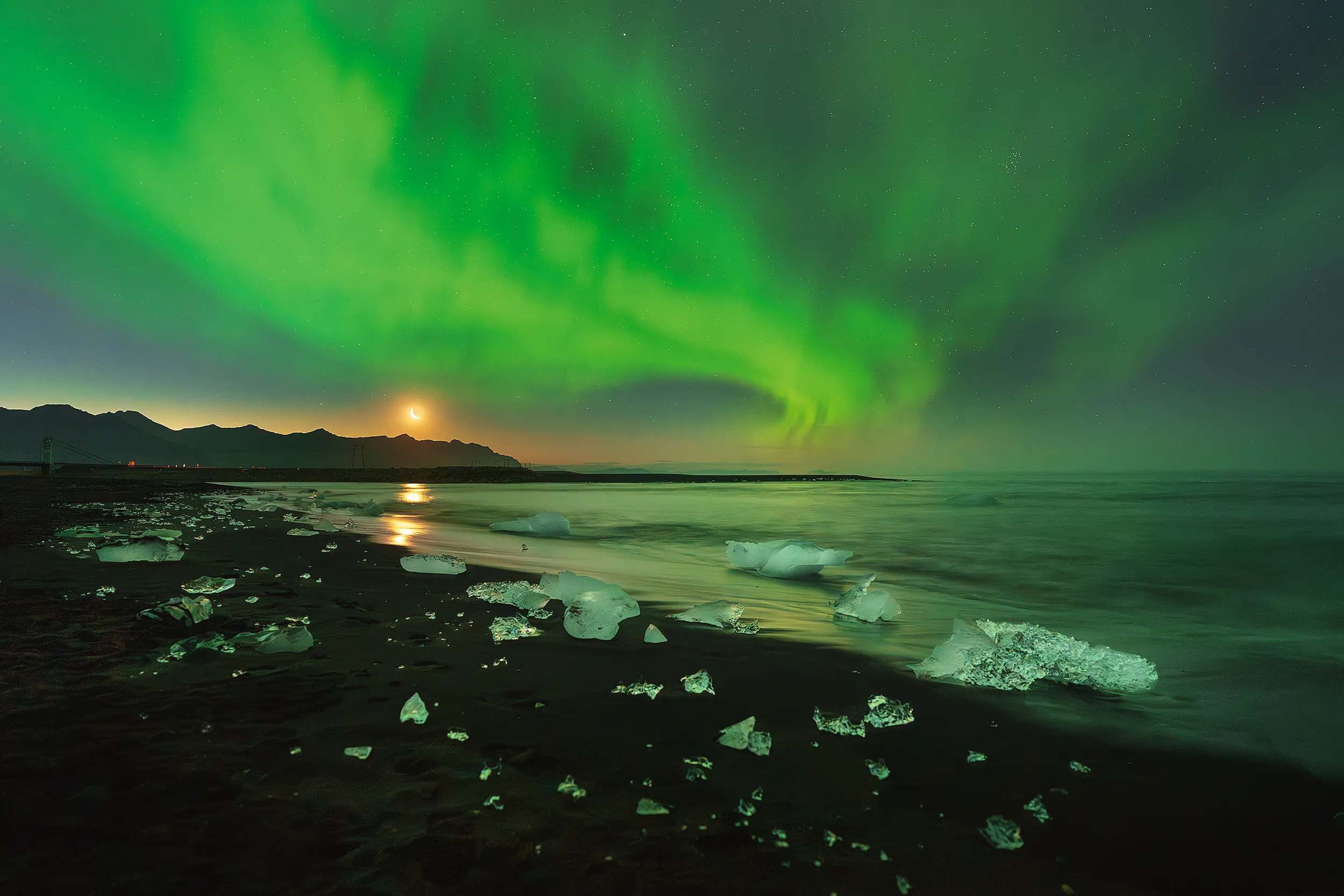 Aurora over a beach scattered with ice, a crescent moon above.