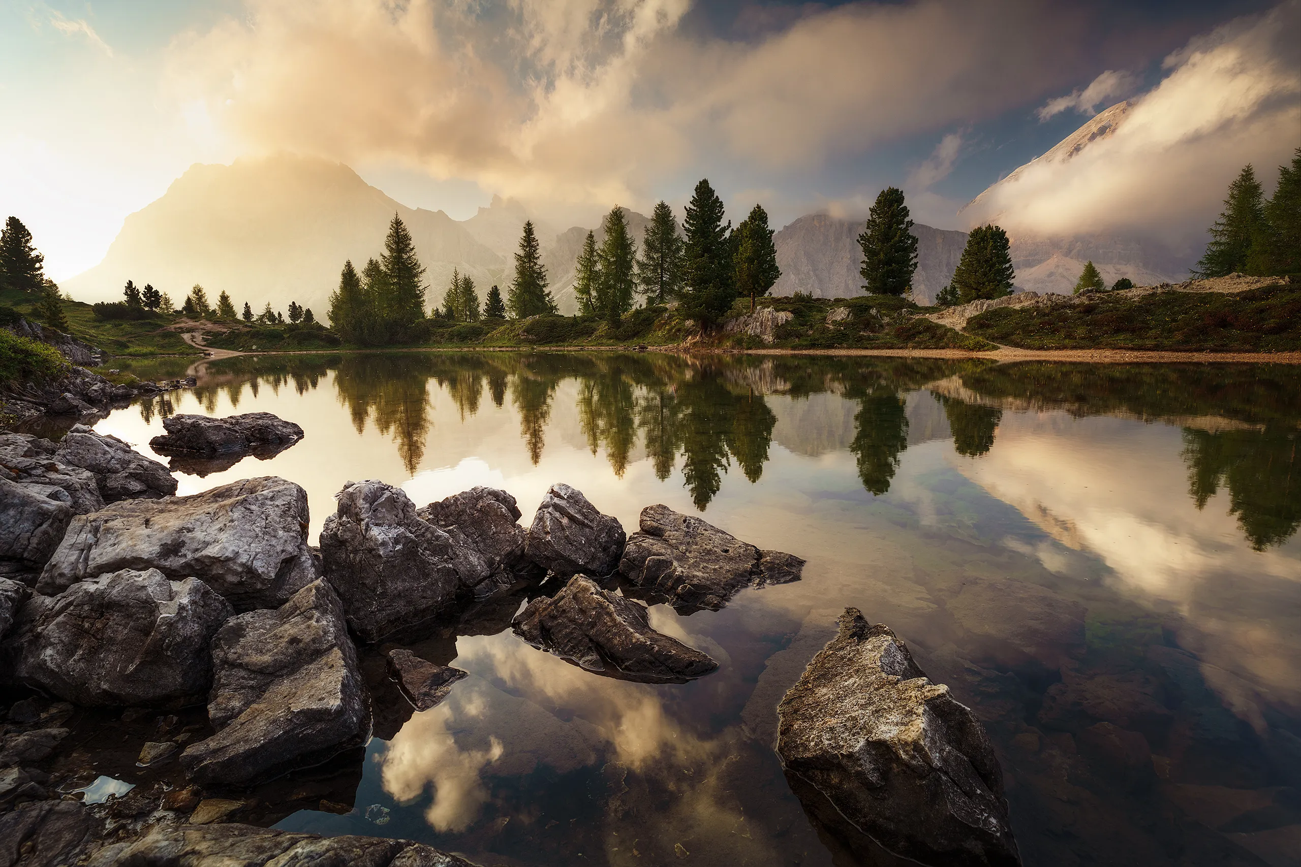 Mountain lake with a rocky shore, calm water reflecting pine trees under a cloudy sky.