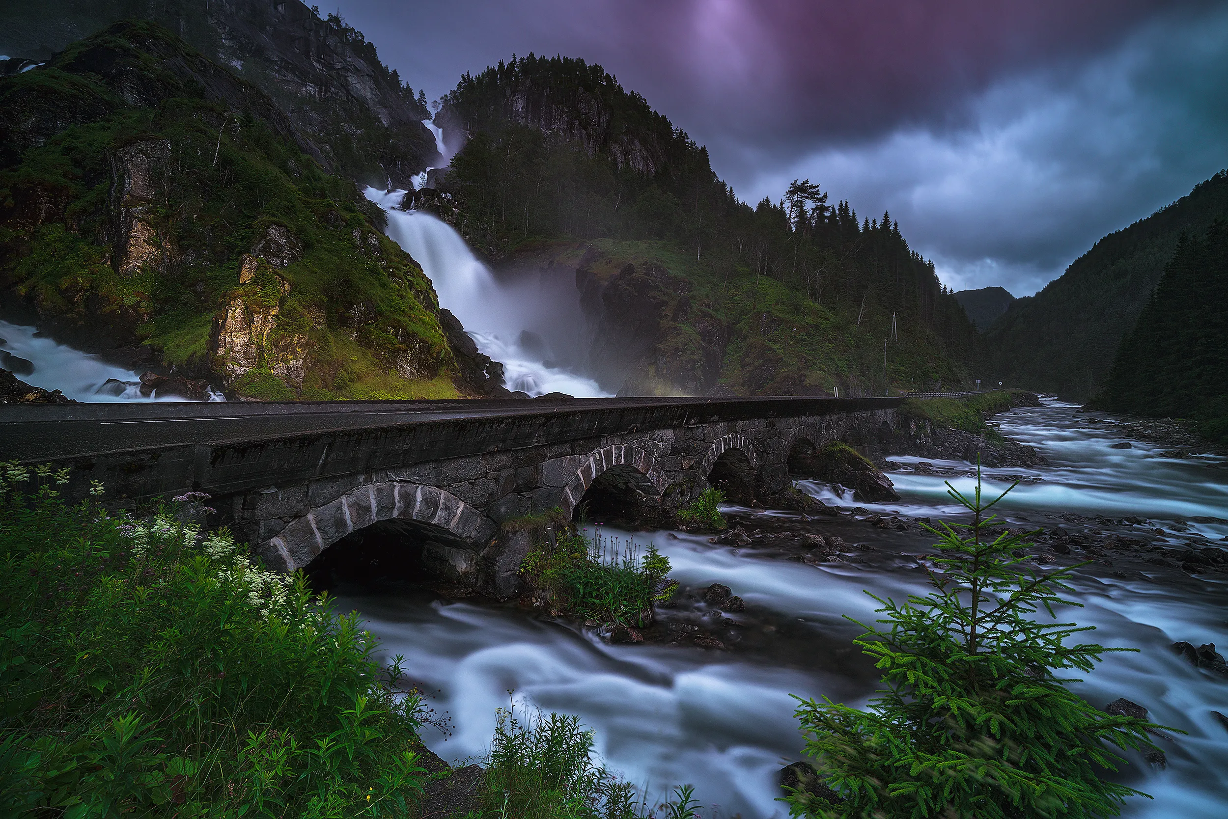A stone bridge over a river, a waterfall behind it under a stormy sky.