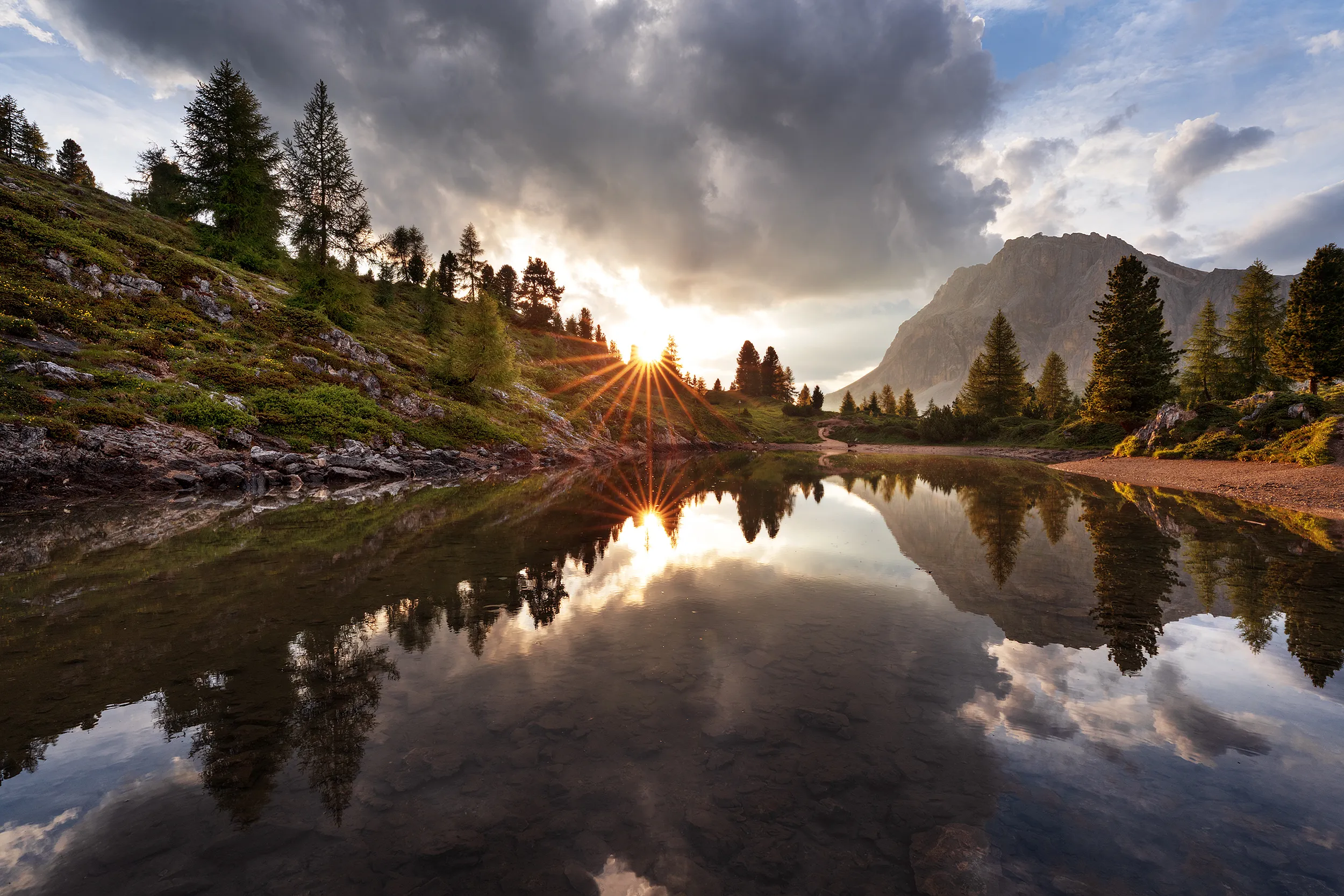 Sun setting behind trees on a hillside, reflected in a calm mountain lake.