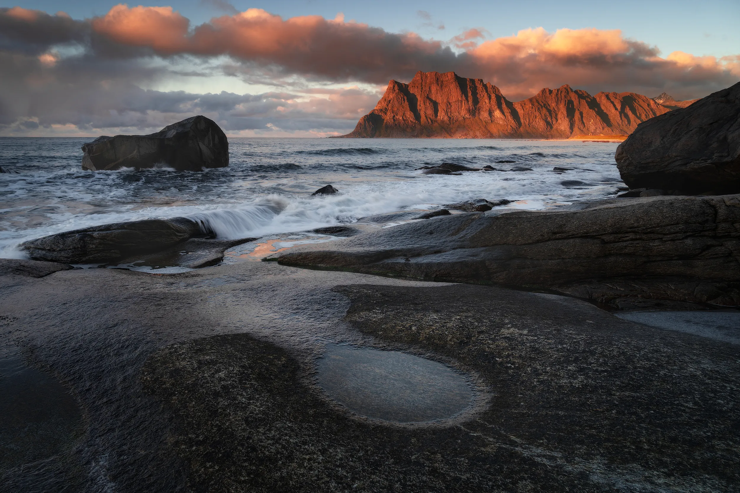 Rocky seashore at sunset with a large distant mountain lit in orange.